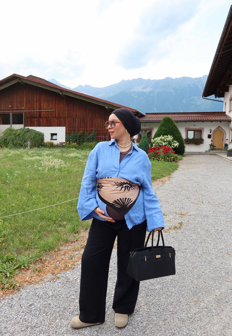 Femme enceinte portant un foulard et des lunettes de soleil, chemise bleue, pantalon noir, tenant un sac à main noir, debout sur un chemin de gravier près de fleurs et d'une maison en bois.