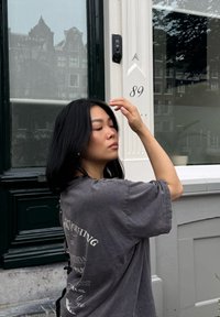 Oversized grijze t-shirt met grafische print op de achterkant, korte mouwen en een ontspannen pasvorm. De achtergrond toont een groene deur met het huisnummer 89.