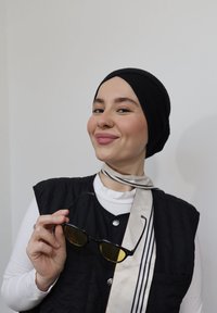 Femme portant un foulard noir et un gilet, tenant des lunettes de soleil à monture noire avec des verres jaunes, souriant légèrement devant un mur blanc uni.