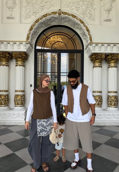 Mujer con hiyab, hombre con barba y niño están dentro de un edificio ornamentado con columnas con detalles dorados y una gran puerta arqueada de cristal.