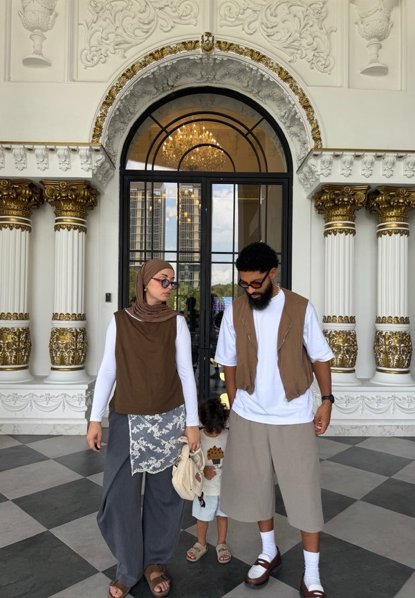 Femeie cu hijab, bărbat cu barbă și copil stau în interiorul unei clădiri ornamentate cu coloane cu accente aurii și o ușă mare de sticlă arcuită.