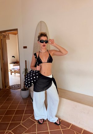 Mujer ajustándose las gafas de sol, con top de bikini negro, pantalones blancos, pareo negro y llevando un bolso negro con lunares, parada en interiores junto a una tabla de surf.