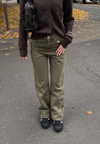 Oliivinvihreät leveälahkeiset housut yhdistettynä tummanruskeaan neuletakkiin, mustaan teksturoituun käsilaukkuun ja mustiin lenkkareihin, joissa valkoiset raidat.