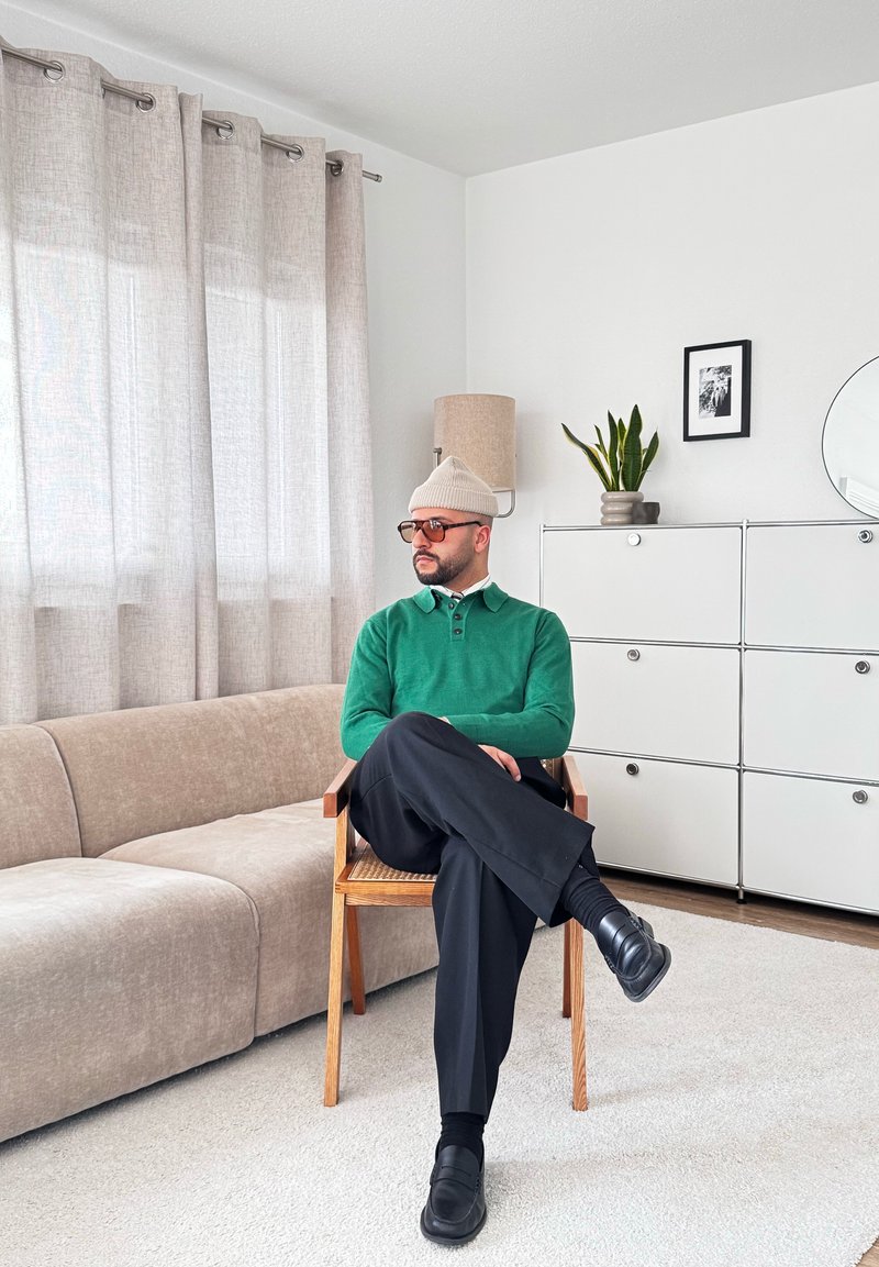 Homme en pull vert et bonnet beige assis jambes croisées sur une chaise en bois dans un salon lumineux avec un sofa beige et des armoires blanches.