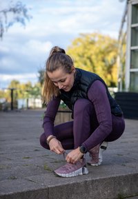 Atletă îmbrăcată cu un vest negru peste un tricou lung violet și colanți, legându-și pantofii de alergare gri și roz pe o suprafață de piatră.