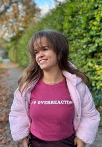 Maglietta bordeaux con testo bianco che dice "I ❤️ OVERREACTING," abbinata a un giubbotto puffoso rosa chiaro. Sfondo naturale con vegetazione.