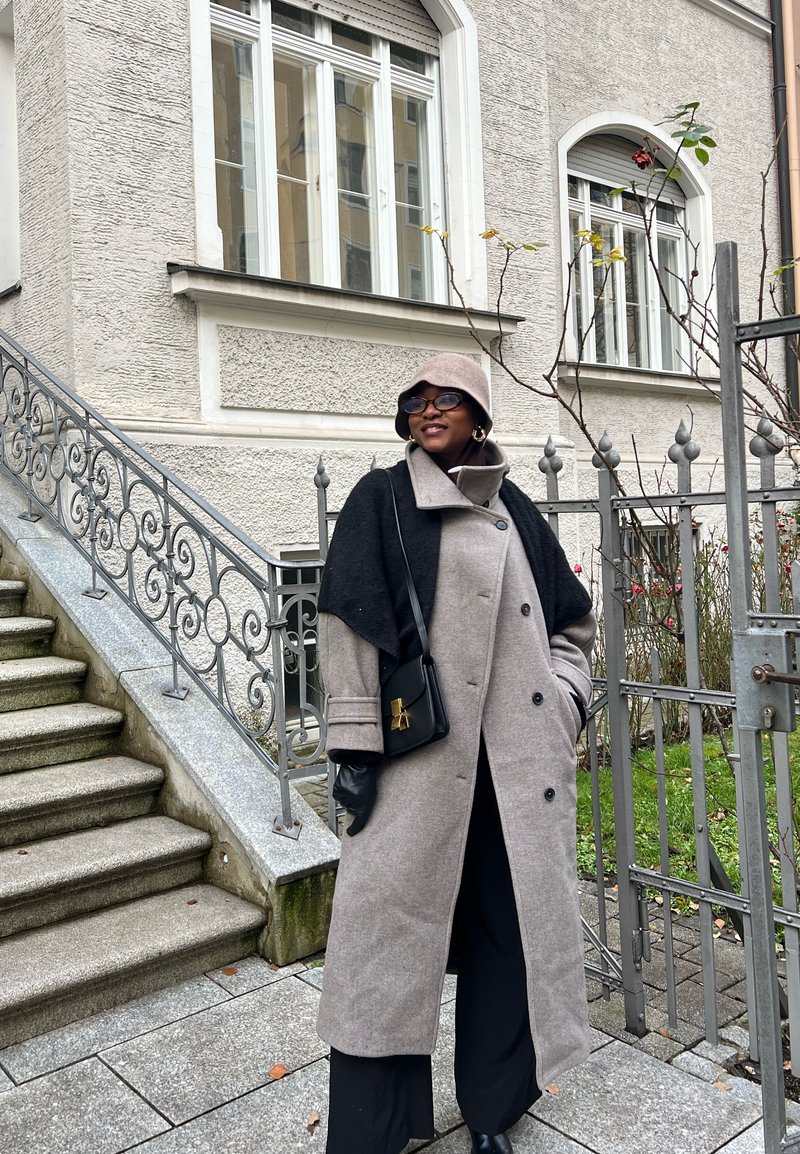 Femme portant un long manteau beige, un châle noir, un chapeau seau, des lunettes et des gants, debout près d'une rambarde ornée et d'une grille devant un bâtiment beige.