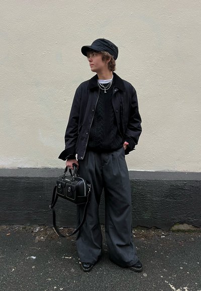 Chaqueta negra sobre un suéter de punto negro, pantalones anchos grises, gorra negra sin estructura, collares de plata y un pequeño bolso negro.