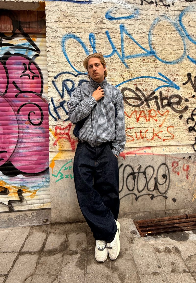 Stribet, langærmet skjorte i grå og hvid, parret med løse, mørk denim bukser. Fodtøjet er lysegrå sneakers. Baggrunden er en væg dækket af graffiti.