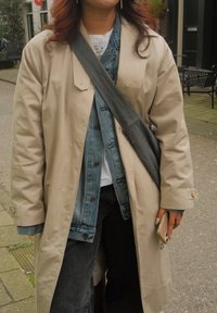 Beige trenchcoat over en denimjakke, kombinert med mørke jeans. Et grått belte krysser over kroppen, og en mobiltelefon holdes i hånden.
