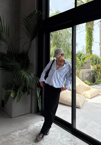 Camisa de manga larga azul claro, pantalones negros y zapatos grises. Ambiente interior con una planta, grandes ventanas y vegetación exterior visible.