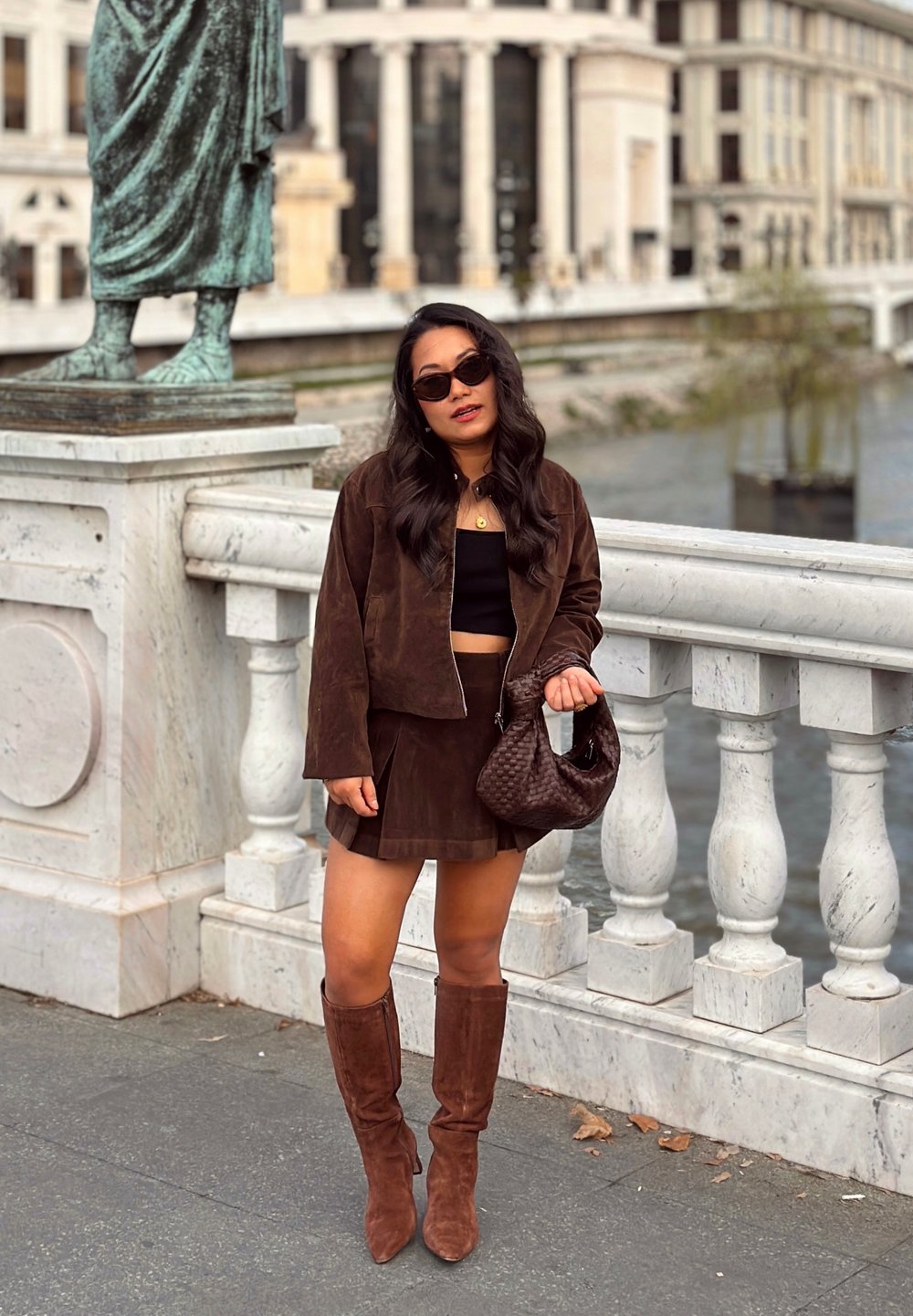Mujer con cabello largo y oscuro, usando gafas de sol, chaqueta marrón, falda y botas, sosteniendo un bolso marrón, de pie junto a un balaustre de piedra cerca del río.
