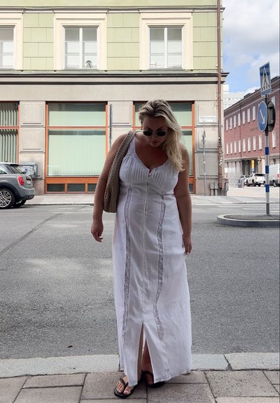 Vestido maxi blanco con rayas horizontales, con detalles de encaje y una abertura en la parte delantera. Combinado con sandalias y un bolso de mimbre.