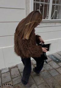 Donna con capelli lunghi che indossa una giacca marrone pelosa e pantaloni scuri, che sistema una borsa nera mentre cammina su una strada lastricata.