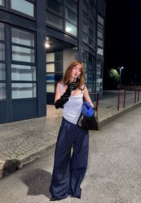 Schwarze lange Handschuhe, graues Tanktop und eine schwarz-blau akzentuierte Tasche. Weit geschnittene navy gestreifte Hosen. Urbane Umgebung mit Glasfenstern.