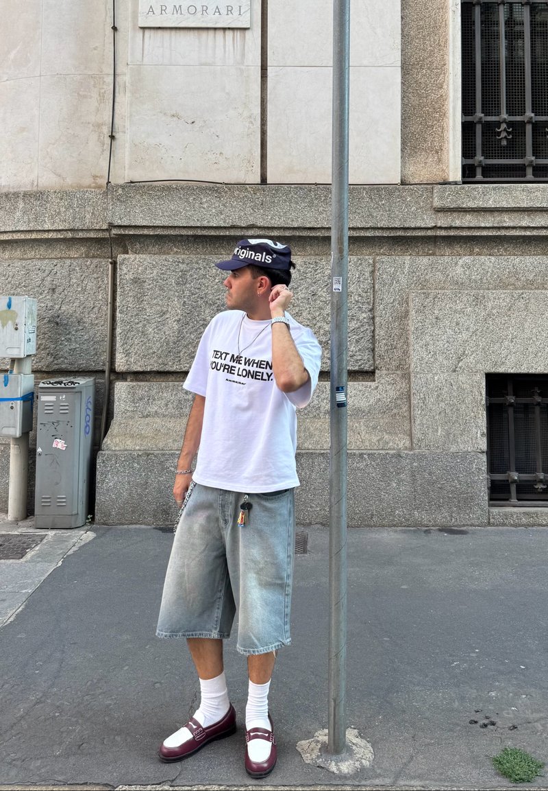 T-shirt blanc avec texte noir, shorts en denim bleu clair, chaussettes blanches et mocassins bordeaux. Les accessoires comprennent une casquette noire et un porte-clés.