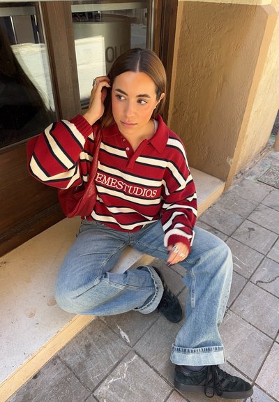 Suéter extragrande a rayas rojas y blancas con cuello de polo, combinado con jeans de un azul claro y zapatillas negras, sentado sobre una superficie de baldosas.