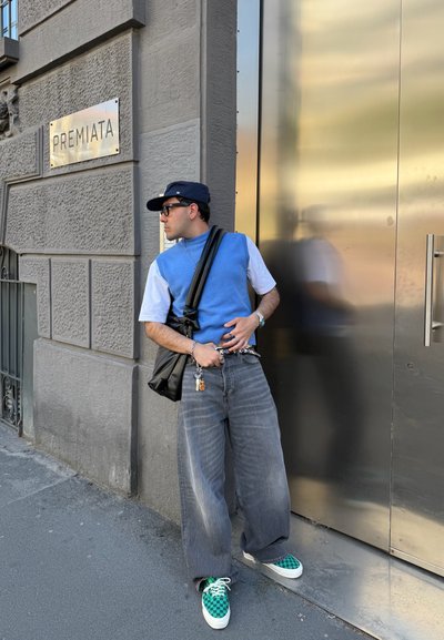Hombre con chaleco azul, camisa blanca, vaqueros grises anchos, zapatillas verdes de cuadros y gorra azul marino apoyado contra una pared fuera de un edificio con puerta metálica.
