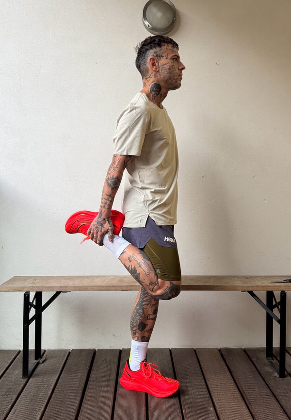 Un hombre tatuado con ropa de correr estira una pierna hacia atrás mientras sostiene una zapatilla de color rojo intenso, junto a un banco de madera.