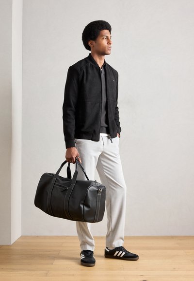 Chaqueta bomber de ante negra sobre un suéter gris, pantalones gris claro y zapatillas negras. Sosteniendo un gran bolso de cuero negro con detalles.
