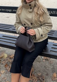 Ruskea nahkalaukku, jossa on jämäkkä muoto, penkillä. Henkilöllä, joka käyttää beigeä trenssitakkia ja mustia lyhytlahkeisia housuja, kädet näkyvissä.