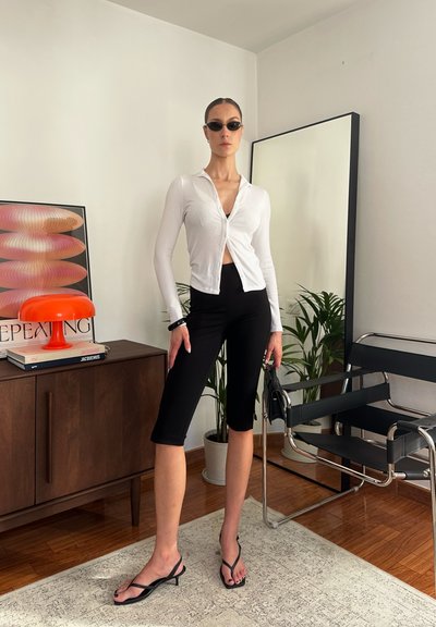 Camisa blanca de manga larga, pantalones cortos negros ajustados, chanclas negras. Habitación con suelo de madera, muebles modernos y una planta. Arte de pared colorido visible.