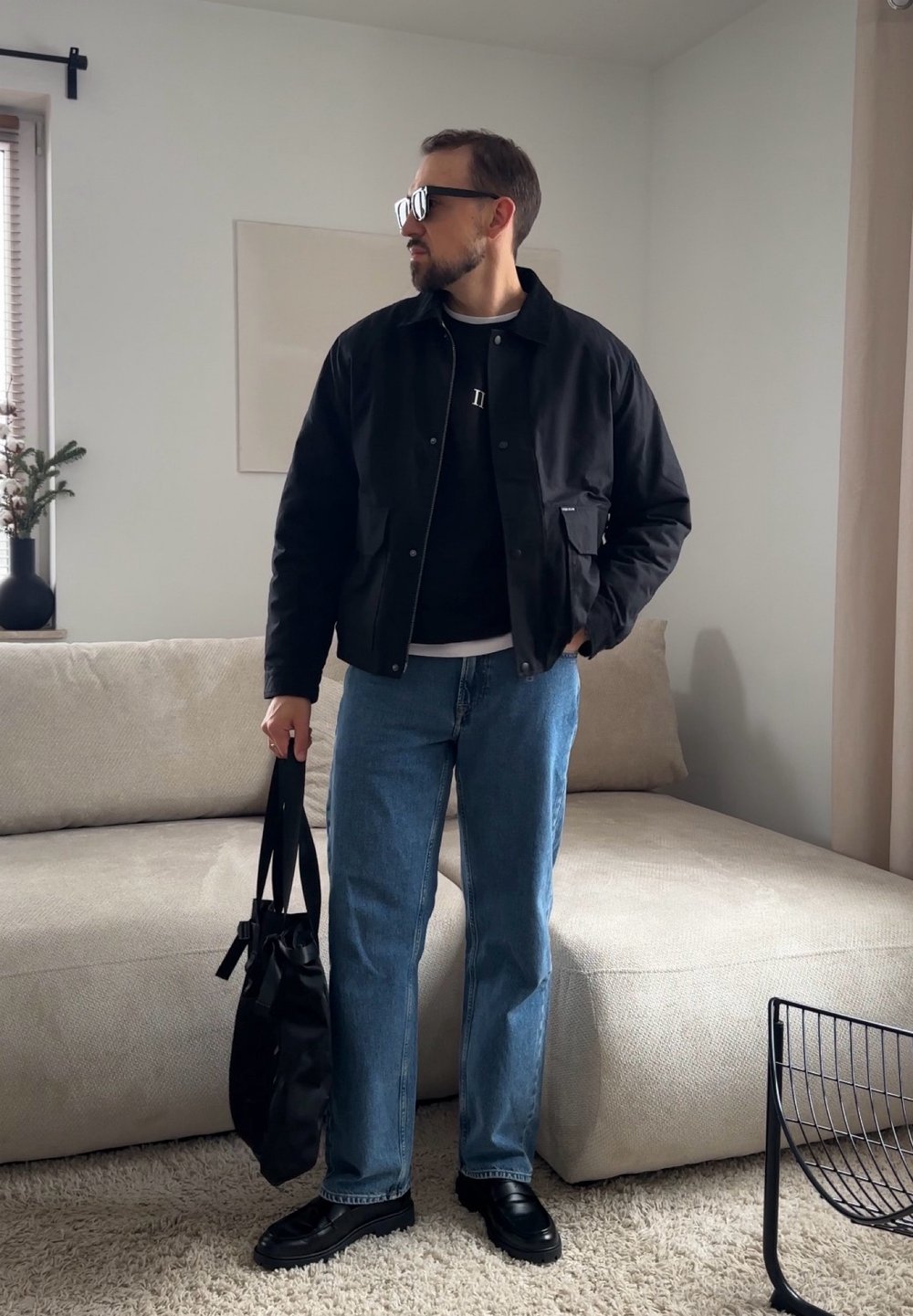Homme debout à l'intérieur portant une veste noire, des lunettes de soleil, un jean bleu, tenant un sac noir, à côté d'un canapé beige et d'une fenêtre avec des stores.