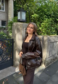 Brązowa skórzana kurtka z zamkiem z przodu i luźnym krojem, noszona na czarnej sukience. Uzupełniona małą brązową torebką z zamszu. Tło z zielonymi liśćmi.