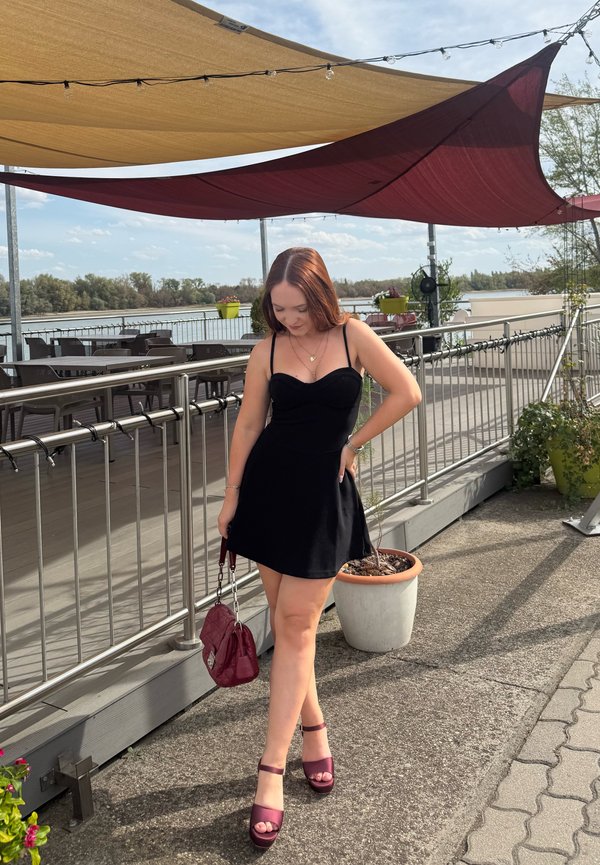 Jeune femme en robe noire tenant un sac à main rouge se tient sur une terrasse au bord de la rivière sous des parasols beige et rouge, regardant vers le bas.