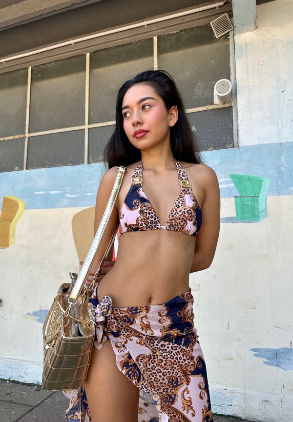 Bikini set featuring a pink and navy leopard print with gold accents, paired with a matching skirt tied at the waist and a metallic gold handbag.