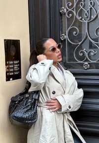 Femme en trench-coat beige et lunettes de soleil ajuste ses cheveux, portant un grand sac noir, se tenant à côté d'une porte noire ornée fermée avec un panneau d'interphone.