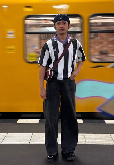 Un hombre joven vestido con una camiseta de rayas en blanco y negro y pantalones negros está de pie en la plataforma del metro con un tren amarillo pasando detrás de él.