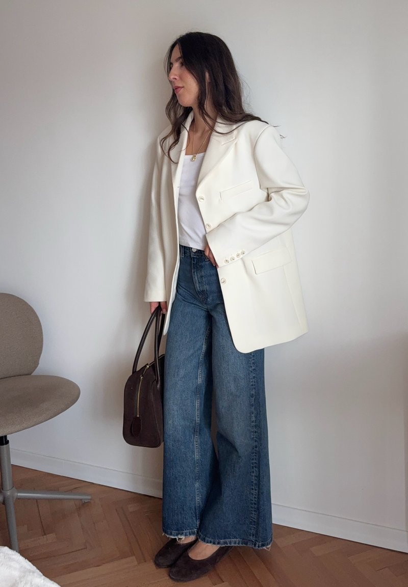 Weißes tailliertes Blazer, weißes Oberteil, weite blaue Jeans, braune Handtasche mit Reißverschluss und dunkelbraune Ballerinas; glatte Stoffe und strukturiertes Design.