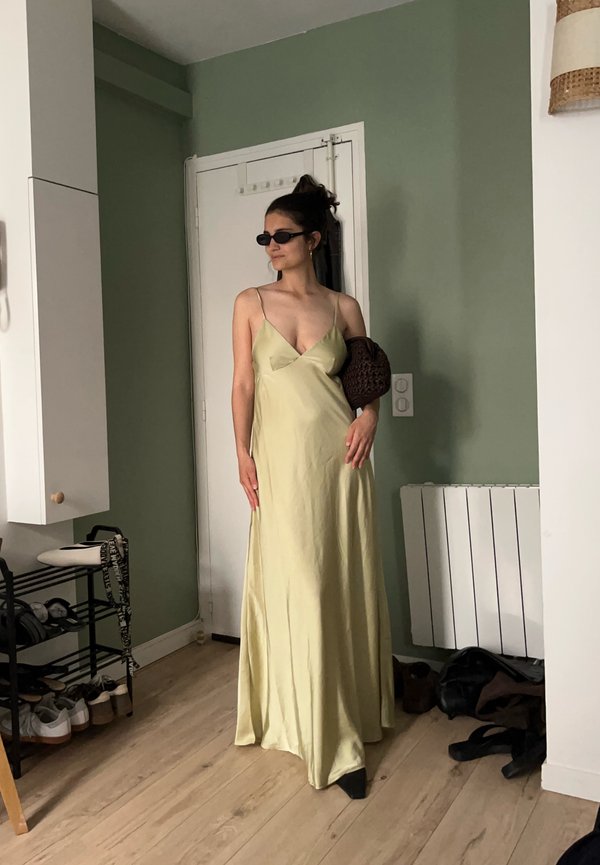 Mujer con un vestido lencero de satén amarillo pálido y gafas de sol negras está de pie en una habitación con paredes verdes sosteniendo un bolso marrón tejido bajo el brazo.