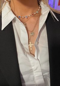 Zlatne ogrlice sa slojevima s privjeskom u obliku srca, križem i choker ogrlicom s višebojnim perlama, nose se preko bijele košulje s ovratnikom.