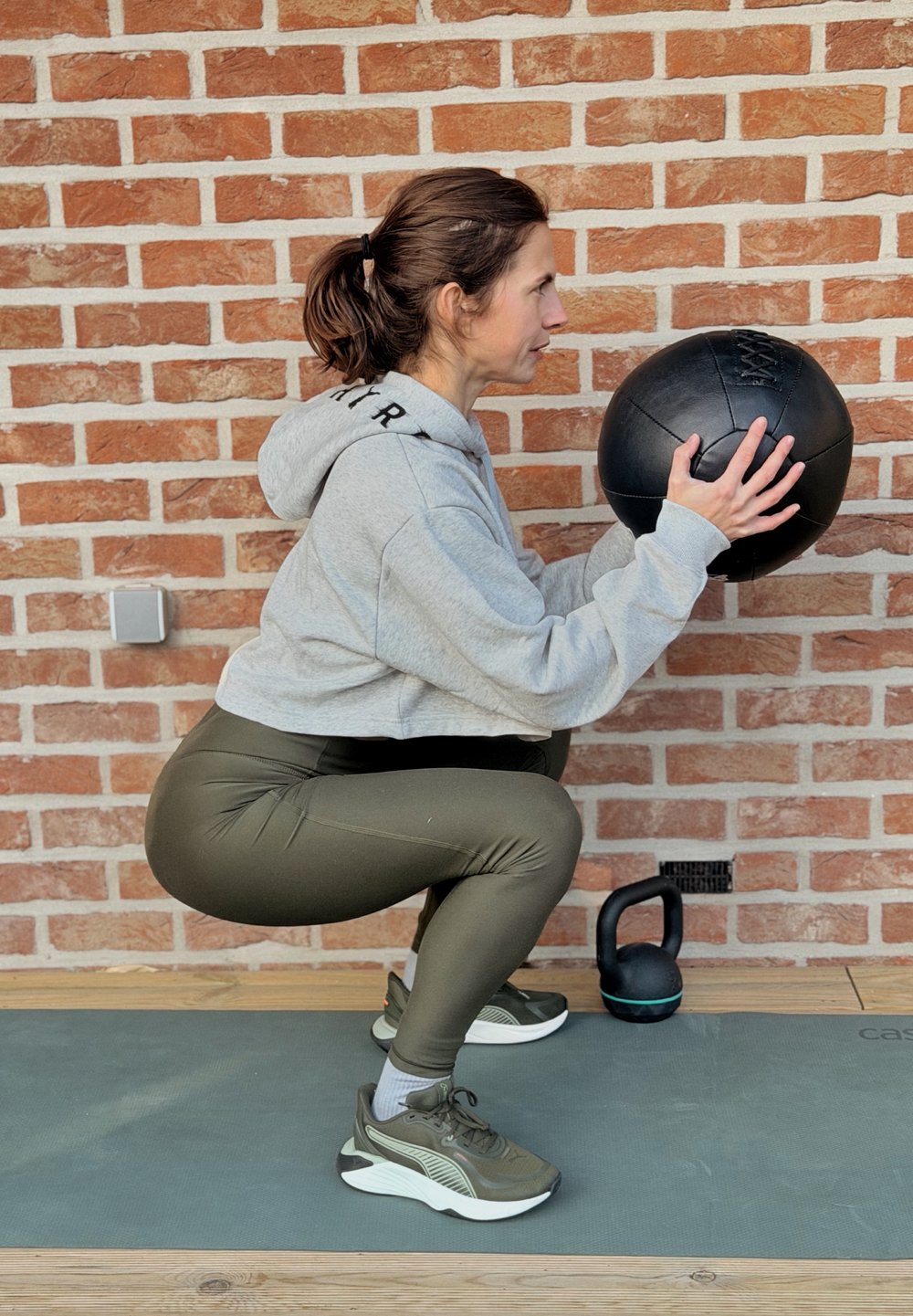 Femme en position accroupie sur un tapis d'exercice tenant un ballon médicinal noir, avec une kettlebell posée au sol à côté d'un mur en briques.