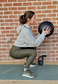 Femme en position accroupie sur un tapis d'exercice tenant un ballon médicinal noir, avec une kettlebell posée au sol à côté d'un mur en briques.