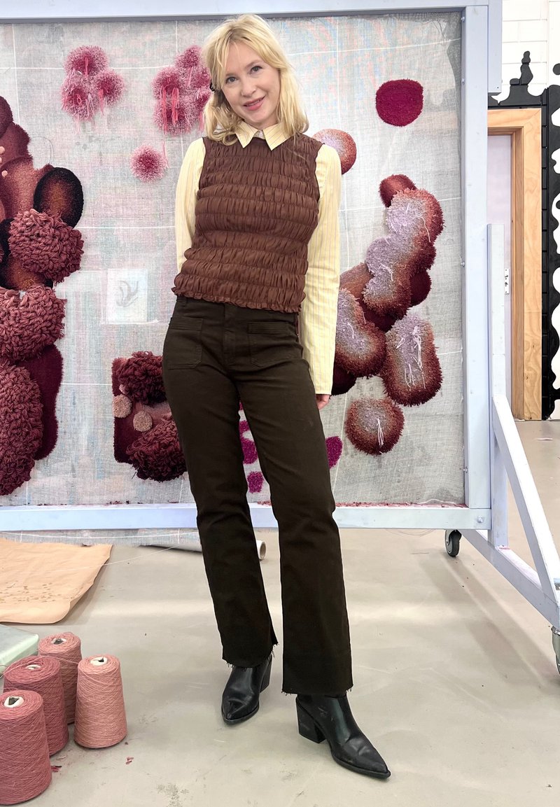Top senza maniche marrone e testurizzato su una camicia colletto giallo chiaro, abbinato a pantaloni a zampa scuri e stivali neri alla caviglia. Lo sfondo presenta arte tessile.