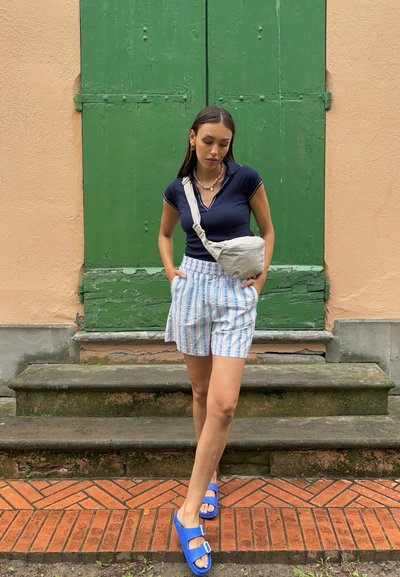 Camisa polo azul marino, pantalones cortos a rayas claros en azul y blanco, sandalias azules, bolso bandolera gris, de pie en escalones de piedra frente a puertas verdes.