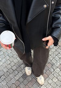Personne portant une veste en cuir noire, un pantalon foncé et des baskets beiges, tenant une tasse de café dans une main et un smartphone dans l'autre, debout sur un pavement en pavés.