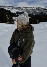 Άτομο με πράσινο φλις μπουφάν και γκρι σκουφί κρατώντας μαύρο μπουφάν, στέκεται σε χιονισμένη πλαγιά με χιονισμένα βουνά και δέντρα στο φόντο.