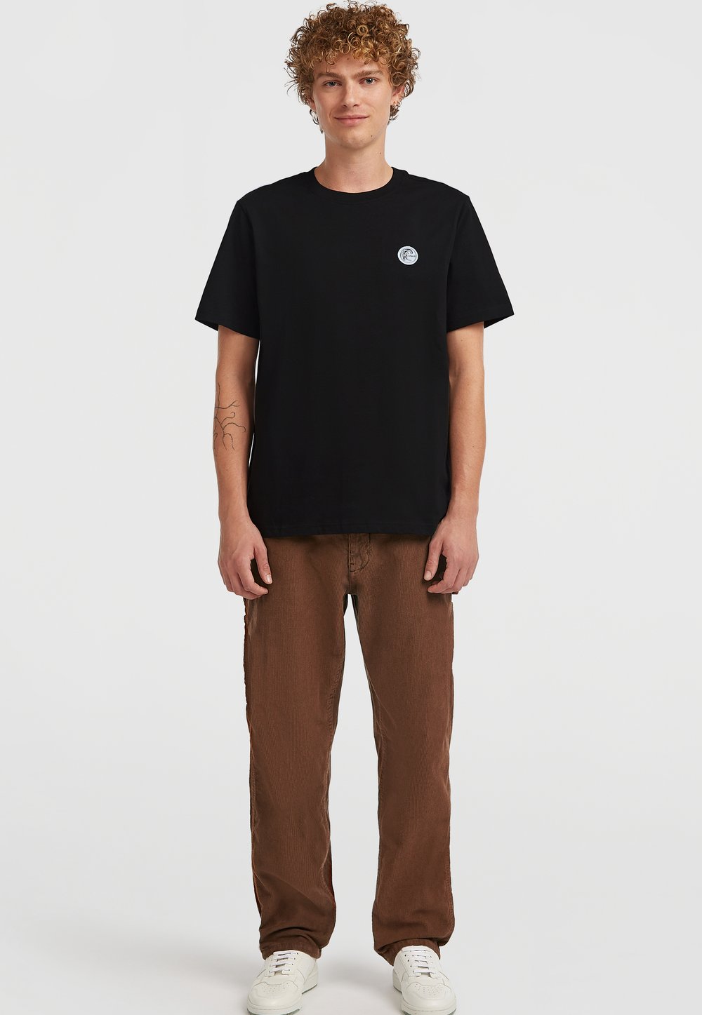 Schwarzes Baumwoll-T-Shirt mit kurzen Ärmeln und Rundhalsausschnitt; verfügt über ein kleines blaues Logo auf der linken Brust. Kombiniert mit braunen, locker sitzenden Hosen.