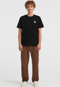 Schwarzes Baumwoll-T-Shirt mit kurzen Ärmeln und Rundhalsausschnitt; verfügt über ein kleines blaues Logo auf der linken Brust. Kombiniert mit braunen, locker sitzenden Hosen.