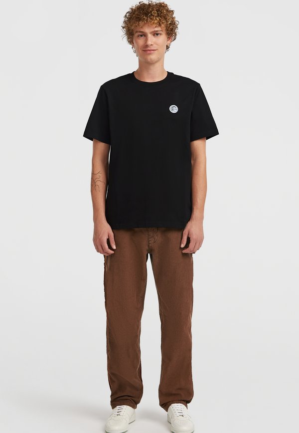 Schwarzes Baumwoll-T-Shirt mit kurzen Ärmeln und Rundhalsausschnitt; verfügt über ein kleines blaues Logo auf der linken Brust. Kombiniert mit braunen, locker sitzenden Hosen.