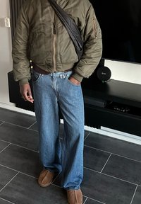 Veste bomber vert olive avec ourlet côtelé, jean large bleu clair, et chaussures en daim marron. Sac bandoulière noir accentue la tenue.