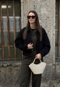 Femme aux cheveux longs portant une veste sombre et des lunettes de soleil, tenant un sac à main blanc, debout devant un mur en béton texturé avec des fenêtres à barreaux.