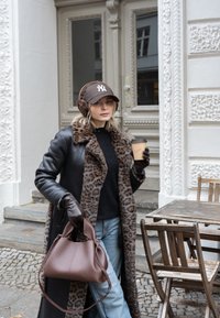 Casaco de couro castanho com colarinho de estampa de leopardo, suéter preto, jeans azuis. Bolsa castanha na mão, segurando uma chávena de café, em um ambiente ao ar livre.