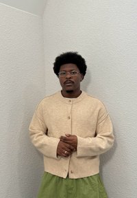 Cardigan en laine beige avec poignets et ourlet côtelés, doté d'un col rond et d'une fermeture à boutons. Associé à une jupe verte. Texture lisse.