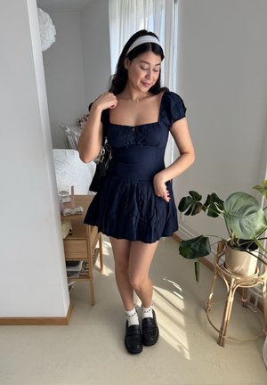 Vestido azul marino con mangas abullonadas, escote cuadrado y falda acampanada. Combinado con mocasines negros y calcetines blancos. Ambiente interior con plantas.