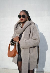 Bežist villasegu oversized-jope suure kraega, mille all on must T-särk ja nahkpüksid, ning käes on pruun nahk-kott.